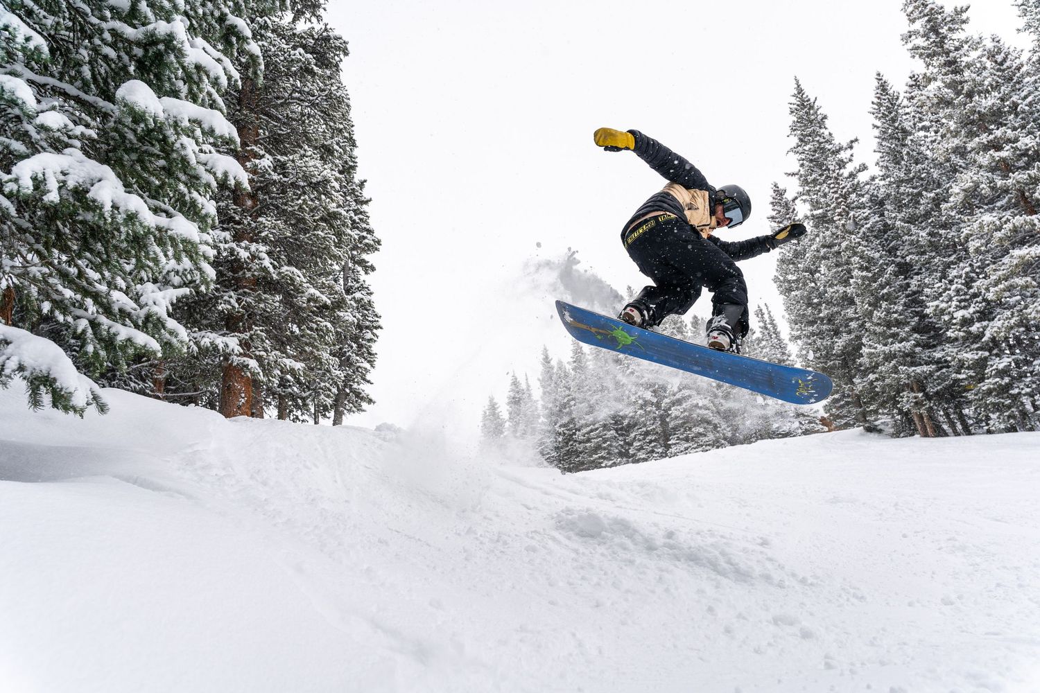 Snowboarder performing a dramatic jump against a backdrop of snow-covered pine trees.