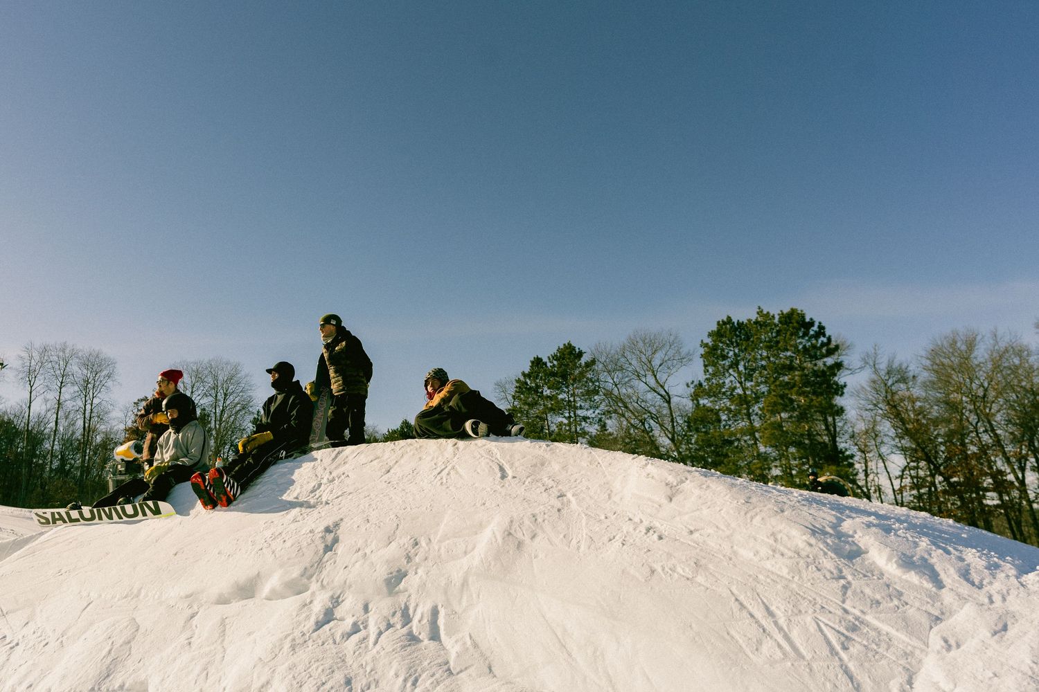 Group of snowboarders standing on a snowy hill against a clear blue sky with trees in the background.