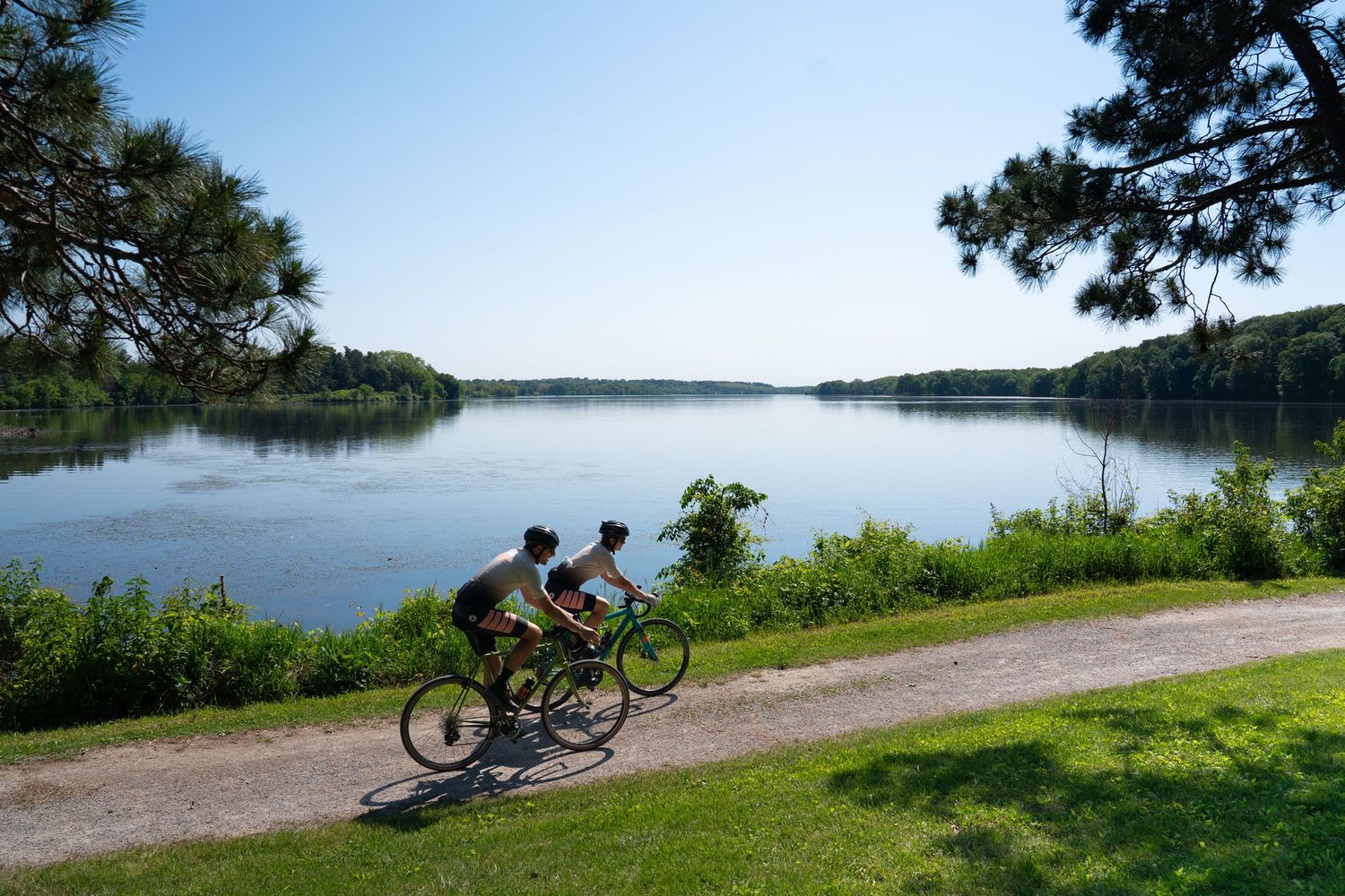 Two cyclists ride along a scenic lakeside gravel path surrounded by lush green grass and trees on a sunny day.