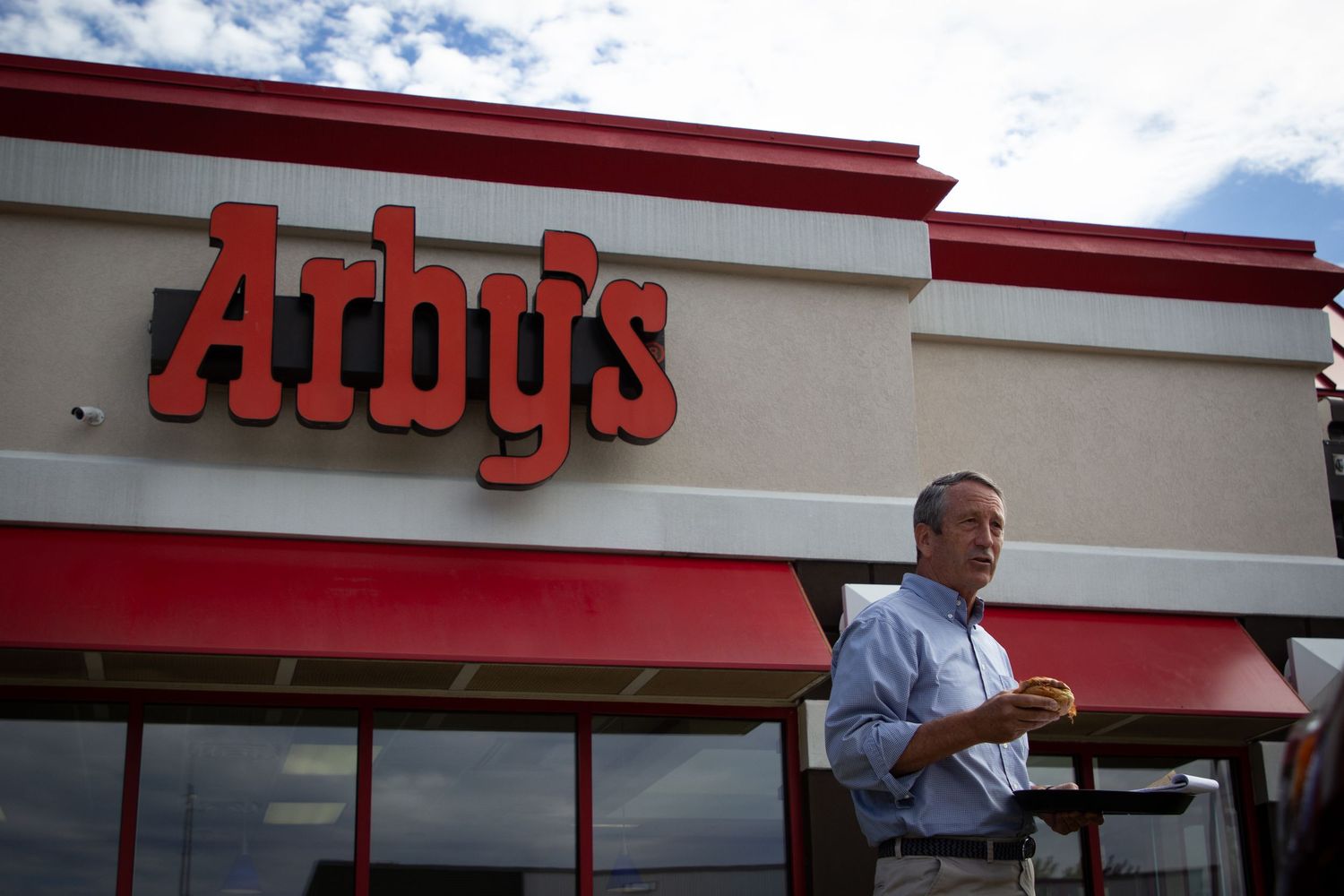 Red and white Arby's restaurant exterior with person standing near entrance under cloudy sky.