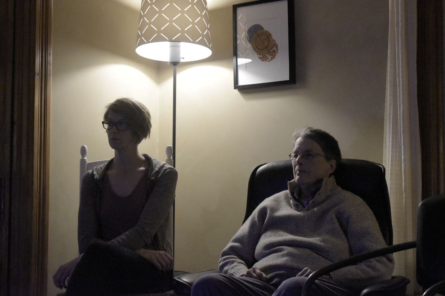 Two people sit in a dimly lit room with a floor lamp and framed artwork on the wall.