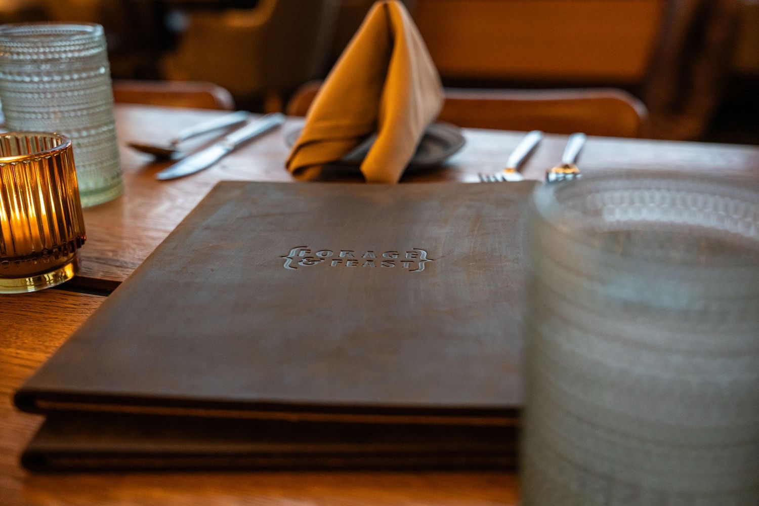 Leather-bound menu and place setting on wooden restaurant table with ambient candlelight.