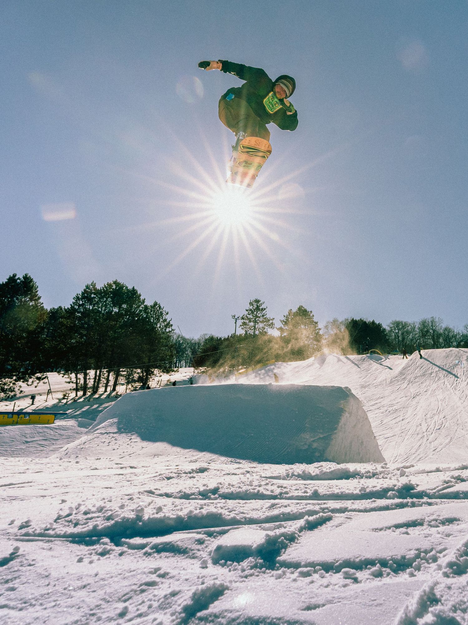 Snowboarder performing a high jump trick at a snow park on a sunny day with bright sunburst and snowy terrain.