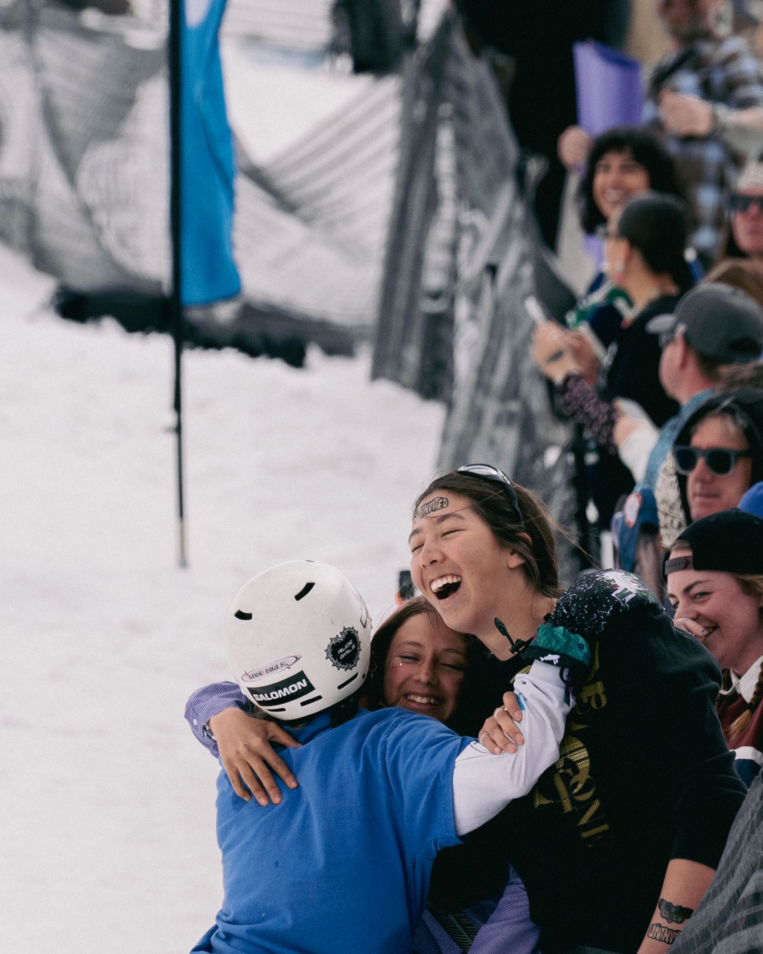 Excited snowboarder celebrates with a hug from a coach near a cheering crowd at a winter competition.