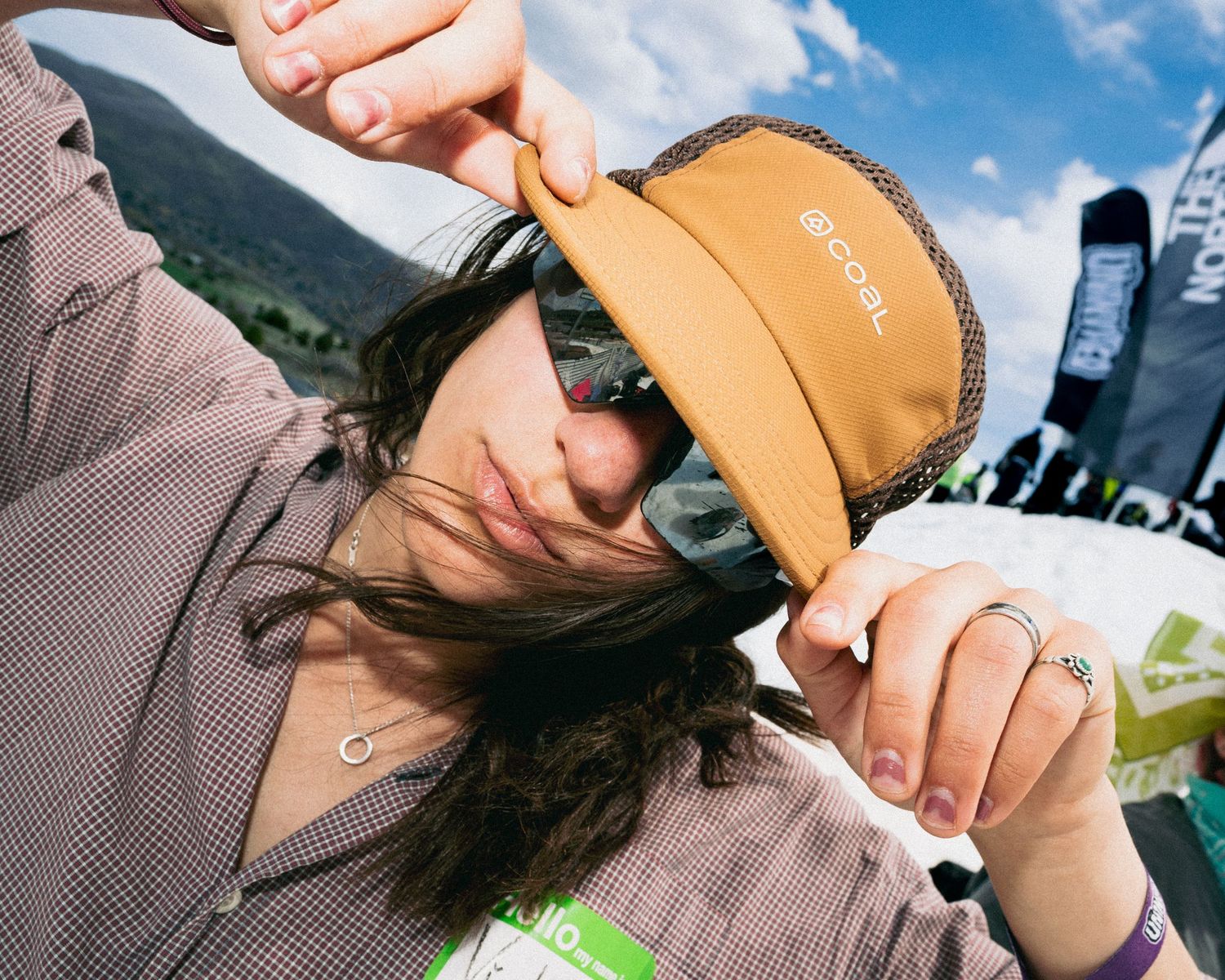 Woman wearing orange cap and sunglasses outdoors at a mountain event on a sunny day.