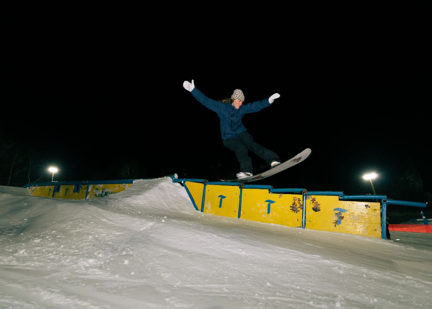 Snowboarder performing a rail grind trick at a night ski park under bright lights on a snowy slope.