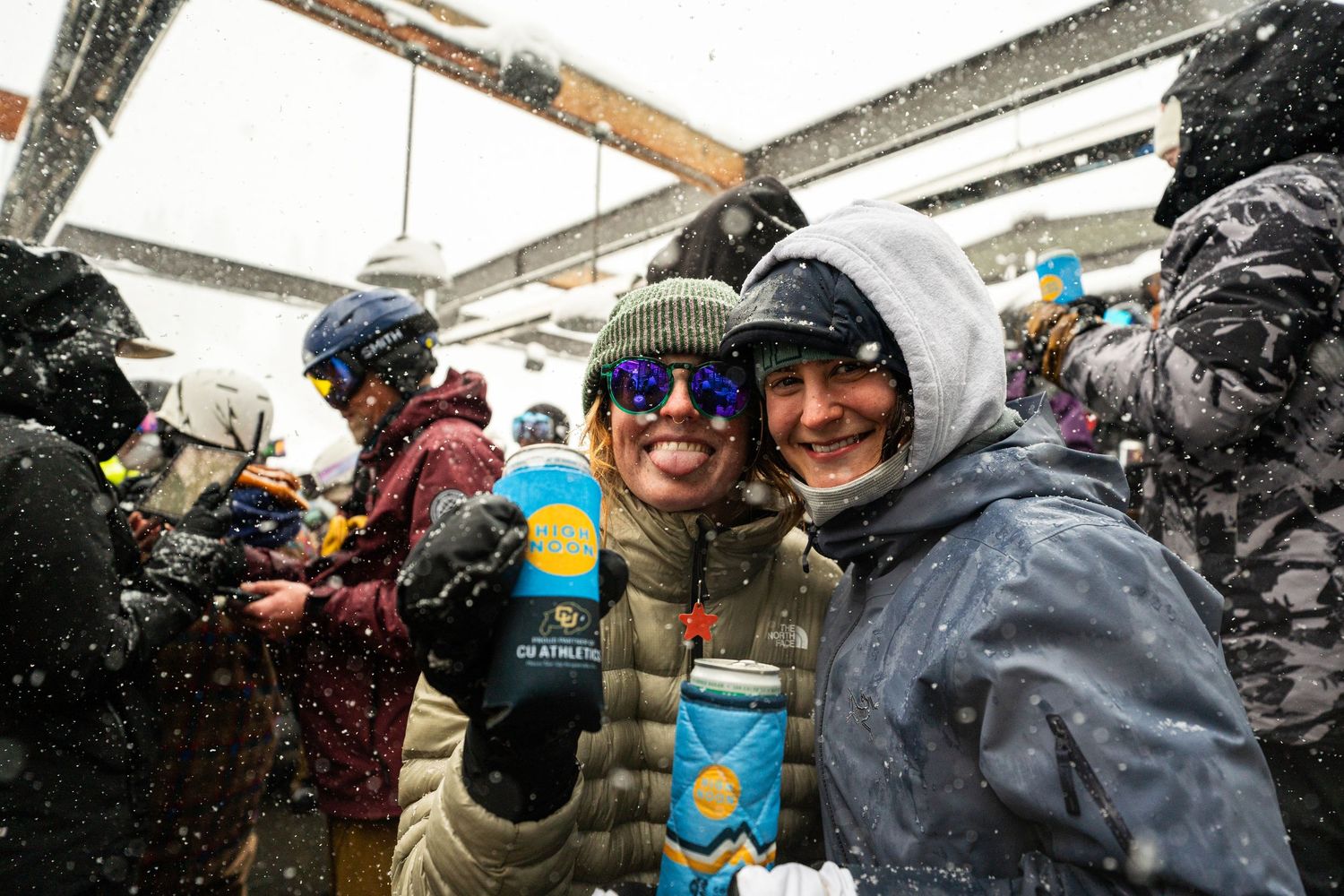 Winter festival attendees in ski gear holding beverages during snowfall.