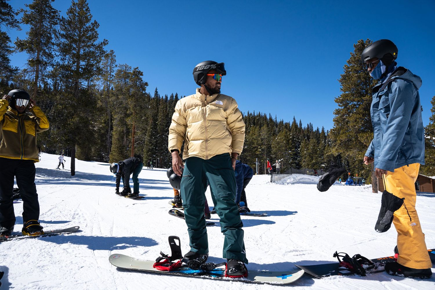 Group of snowboarders standing on snowy slopes with evergreen trees in background on a sunny winter day.