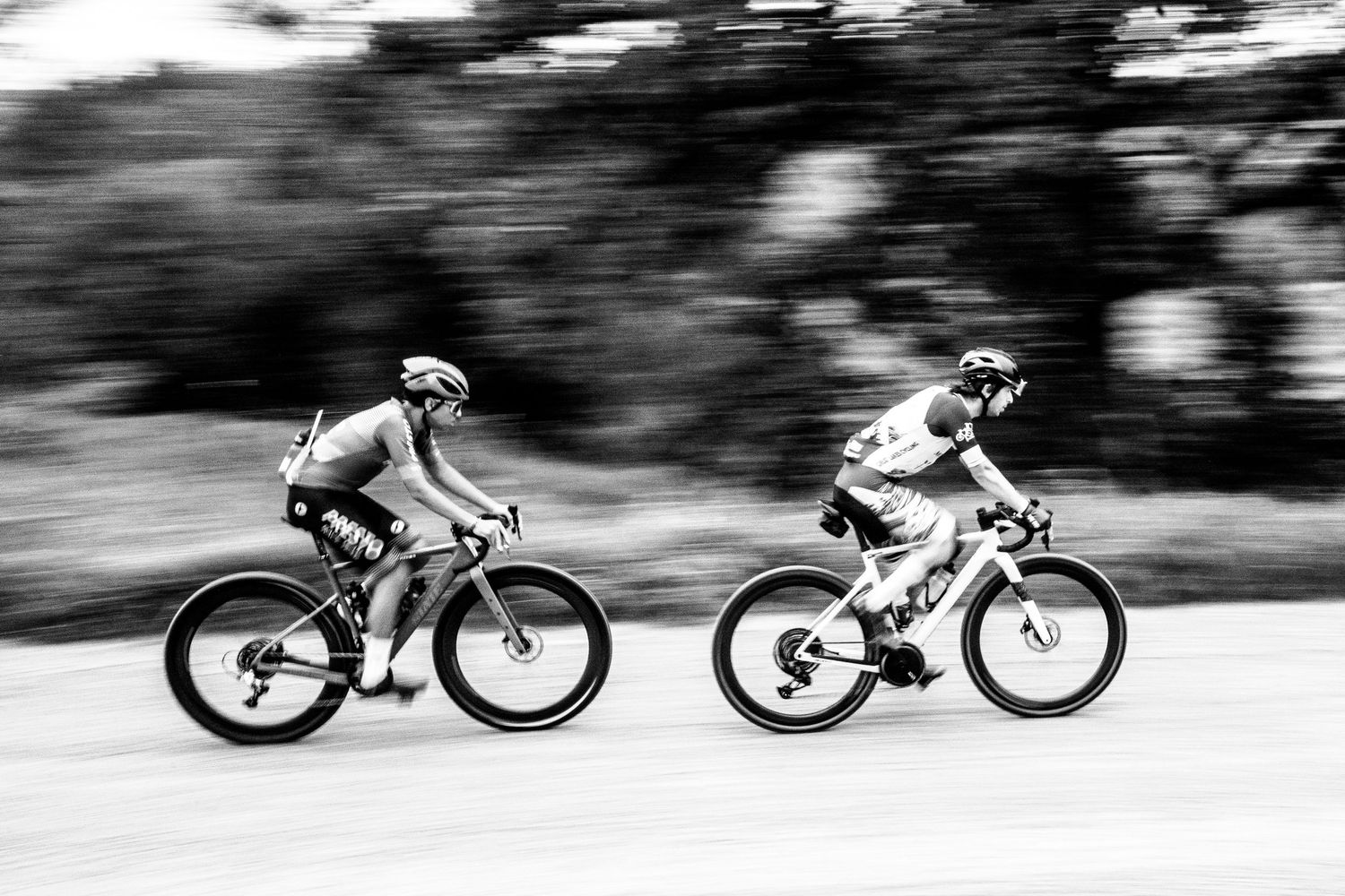 Two cyclists race side by side on road bikes in a dynamic black and white action shot with motion blur background.