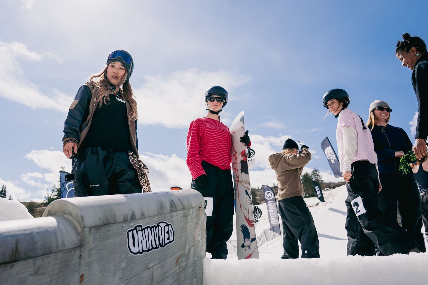 Snowboarders gather on a snowy slope under a bright blue sky, holding boards and wearing winter gear at a ski resort.