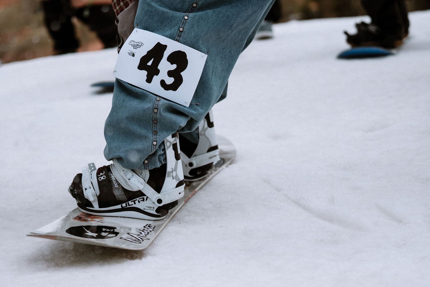 Close-up of snowboarder wearing bib number 43 with bindings on a snowboard at a competition.