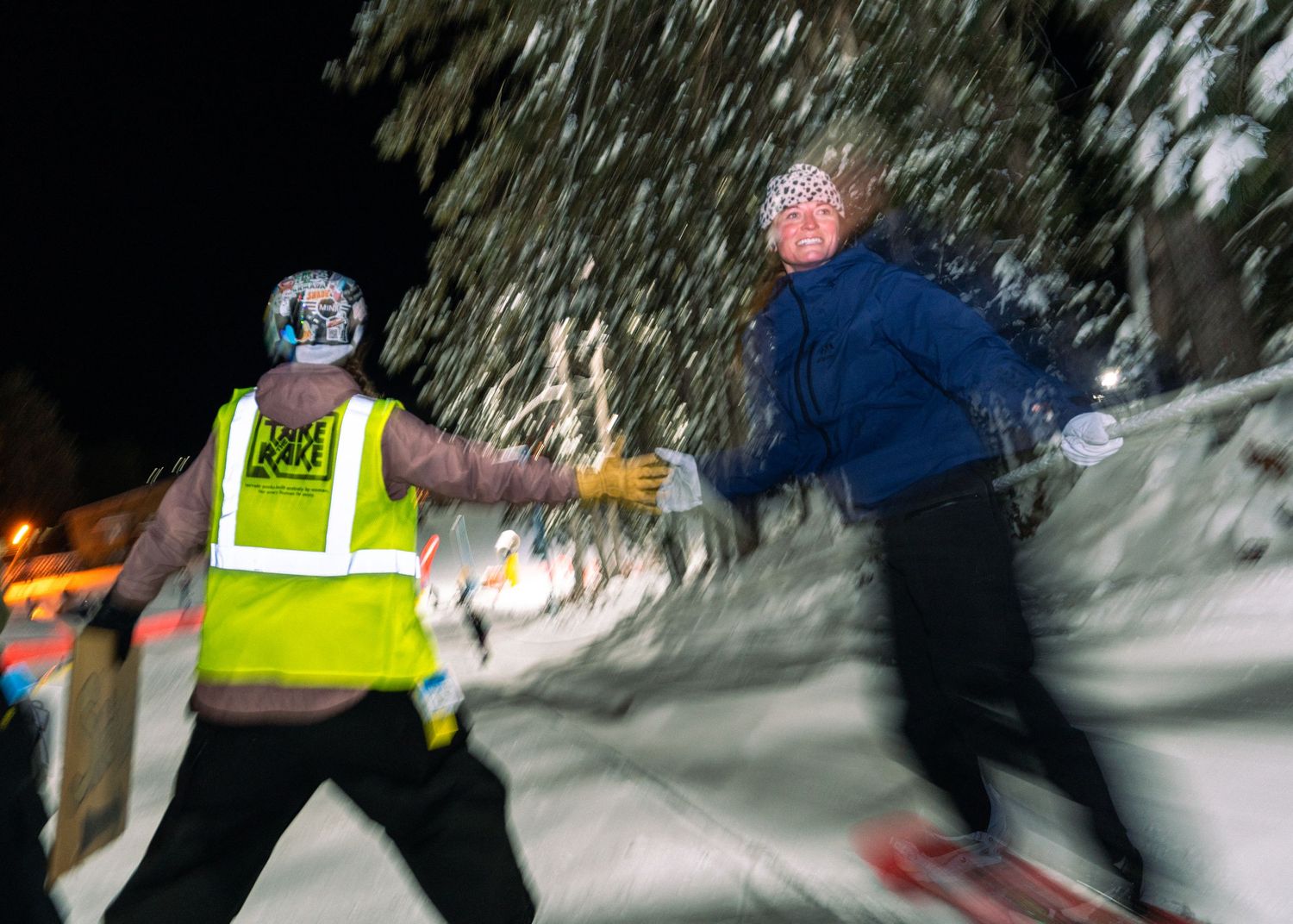 Night ski race runner receives baton handoff from volunteer in yellow safety vest on snowy trail.
