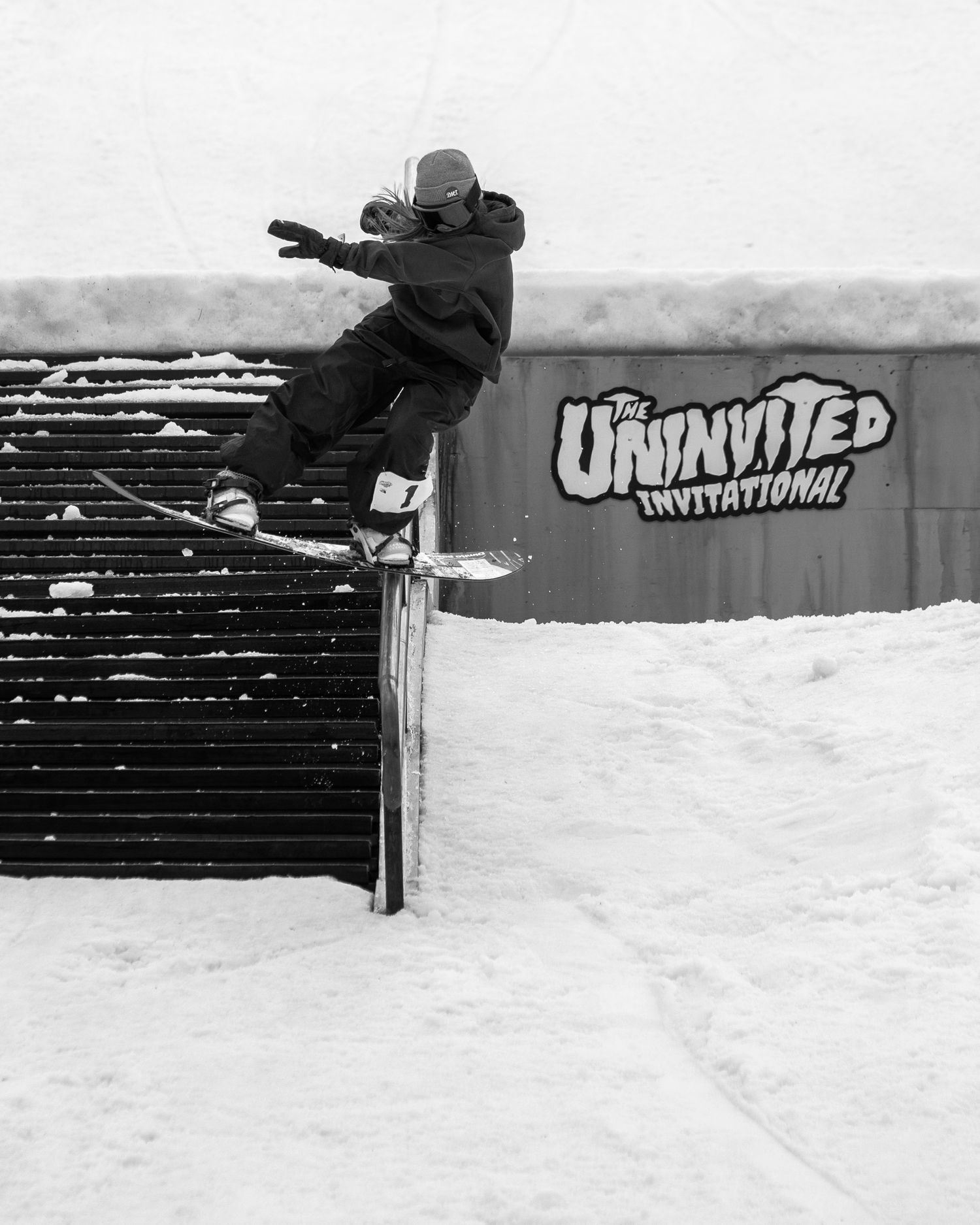 Snowboarder performs a rail grind trick at The Uninvited Invitational contest in snowy conditions, captured in black and white.