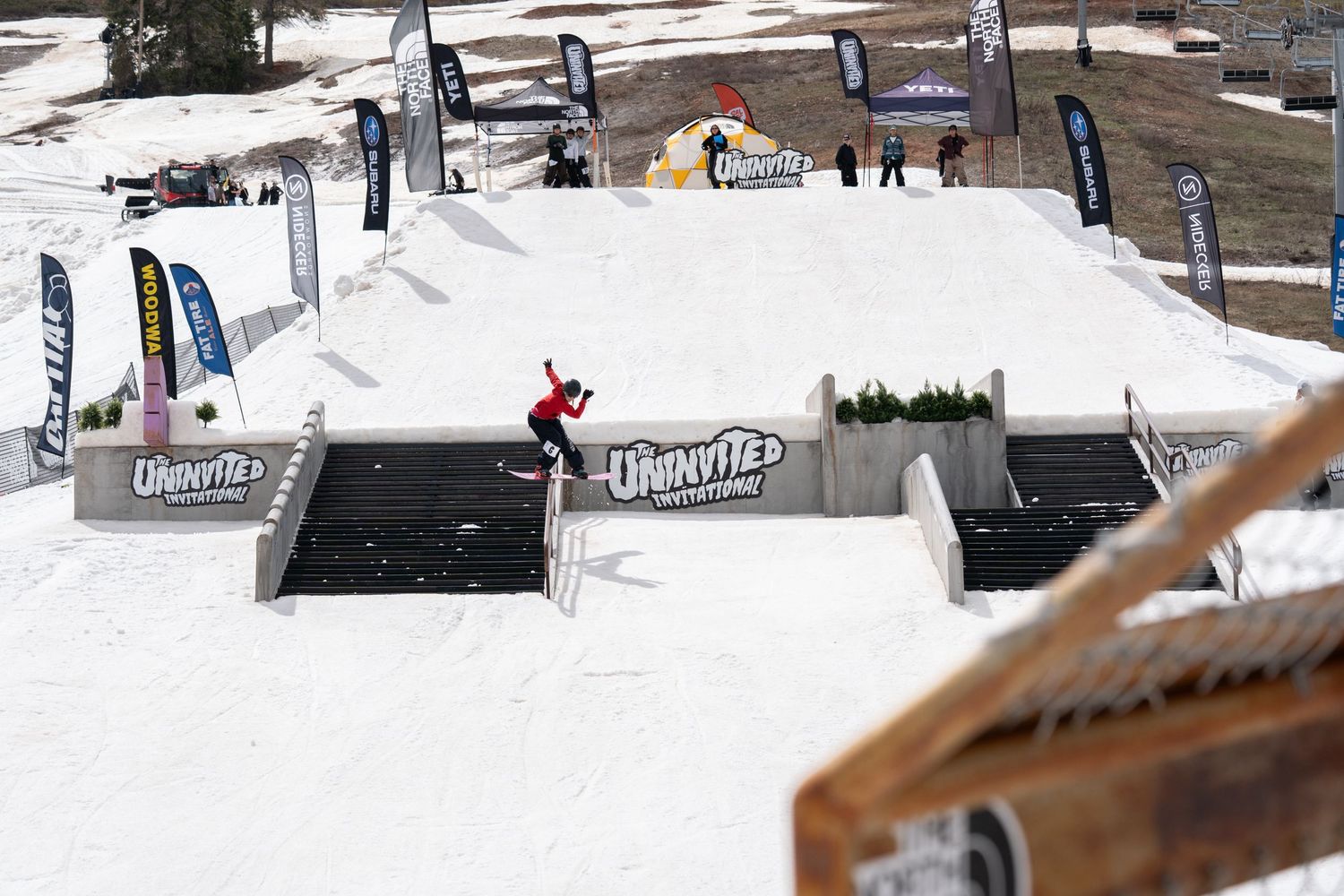 Snowboarder performing a trick on a rail at an outdoor snow park competition with colorful banners and flags.
