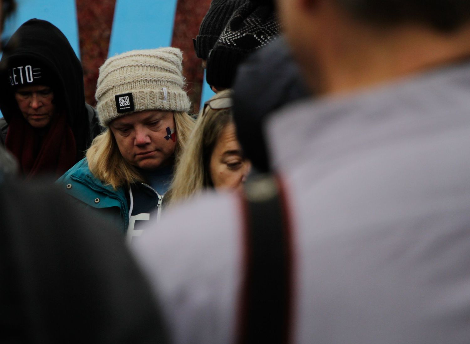Crowd of people wearing winter hats gathered outdoors in muted lighting.
