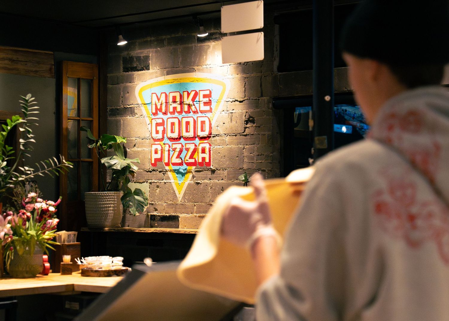 Person stretching pizza dough in front of a neon Make Good Pizza sign on a brick wall.