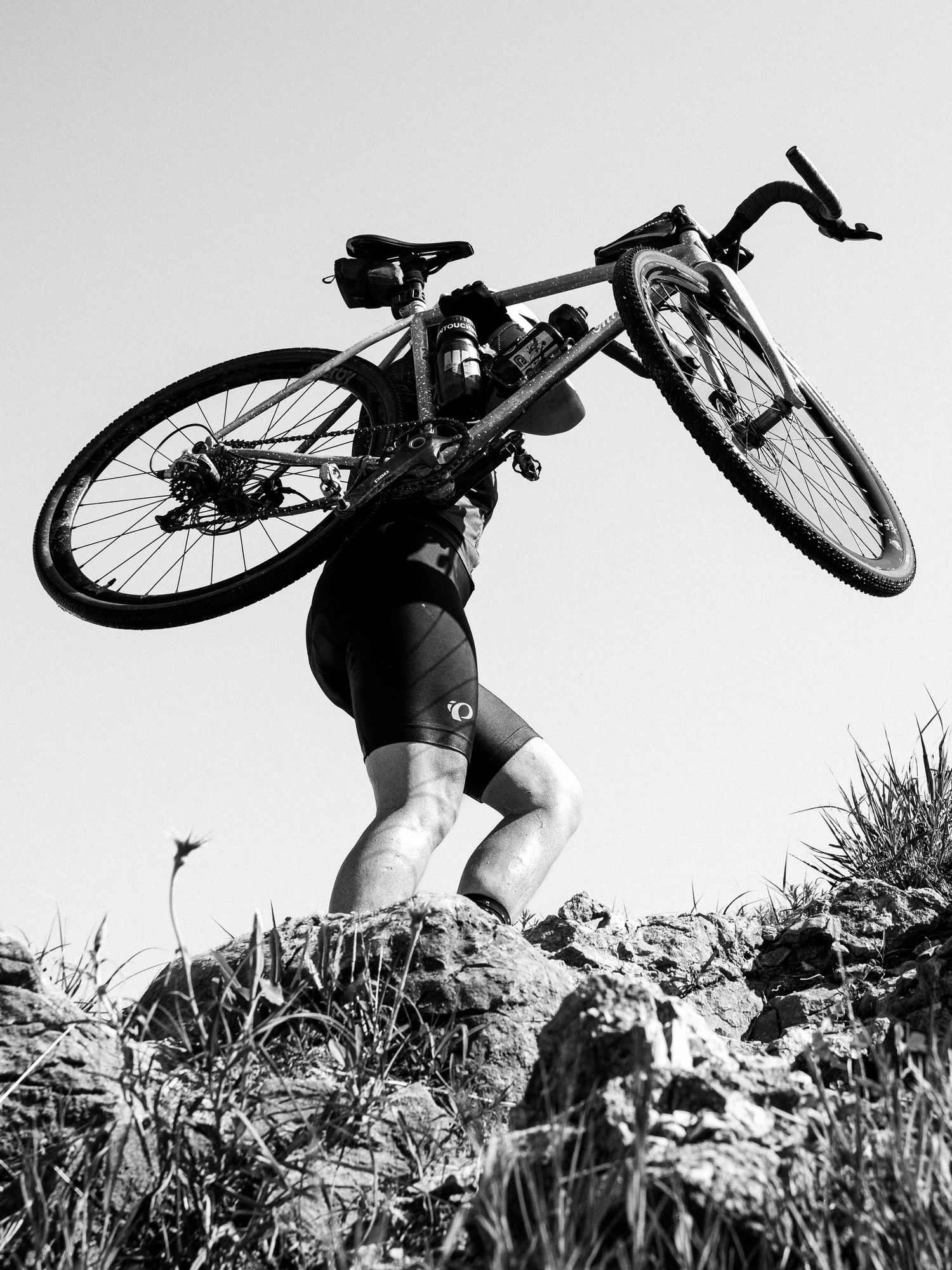 Black and white photo of a cyclist lifting a road bike overhead while standing on rocky terrain outdoors.