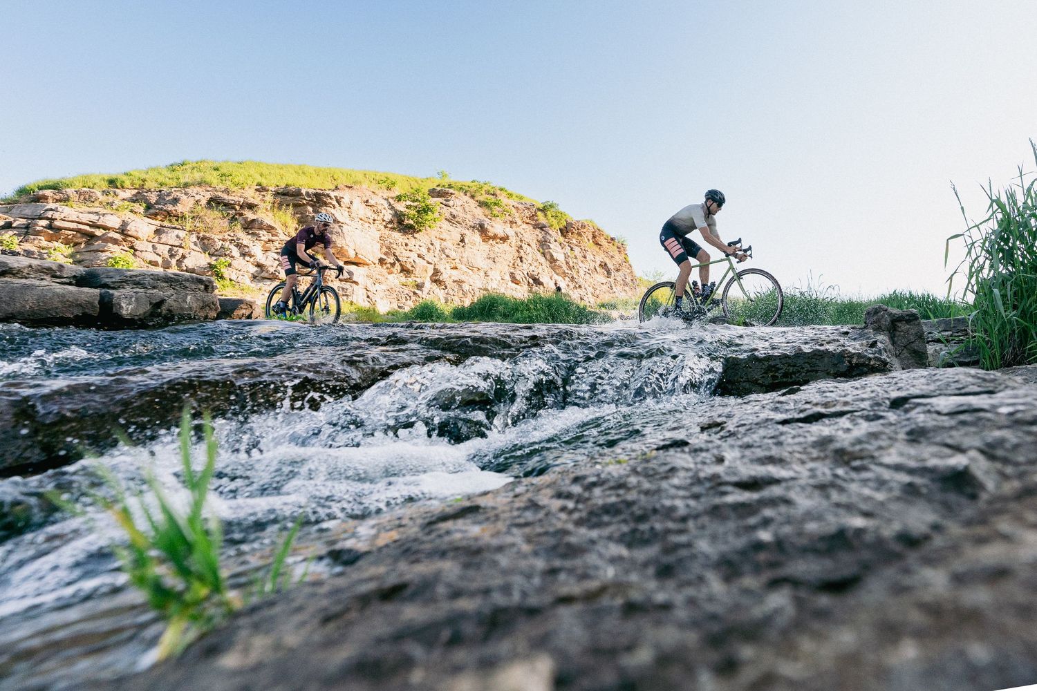 Two cyclists ride mountain bikes along a rocky trail beside a rushing stream with a green hillside backdrop.