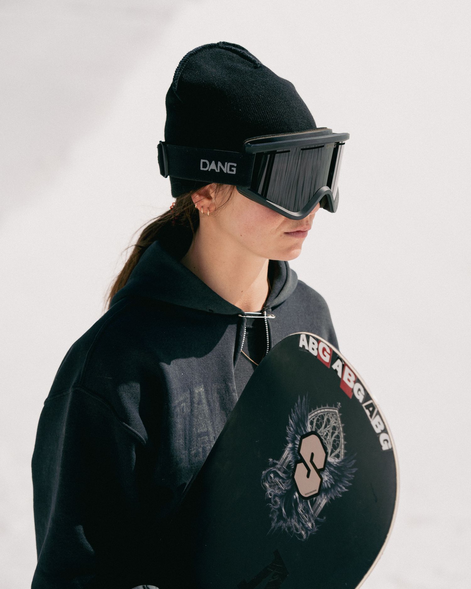 Woman wearing black DANG goggles and beanie holding a snowboard in a studio setting.