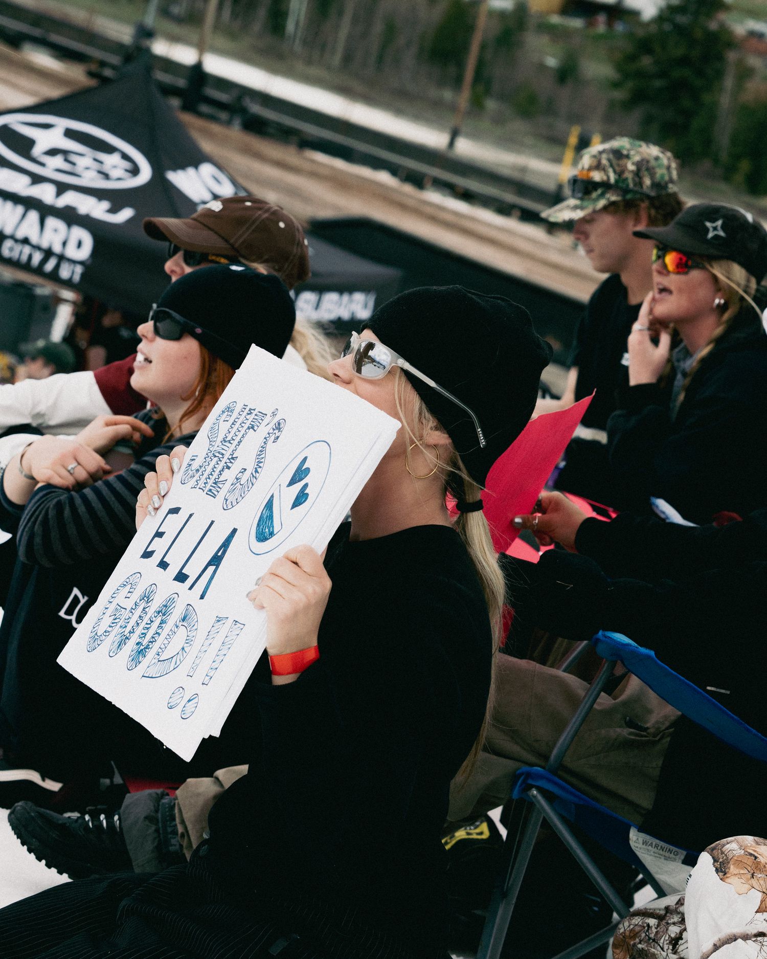 Spectators at a snowboard event, one holding a sign cheering for competitor Ella.