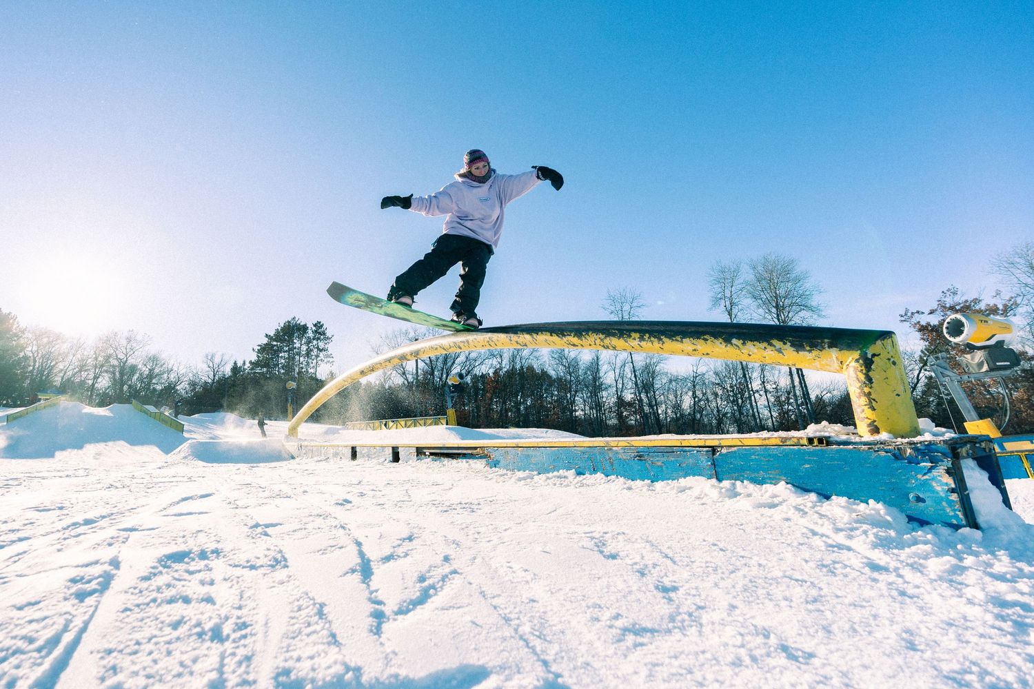 Snowboarder performing a jump trick over a yellow rail feature at a snow park on a sunny winter day.