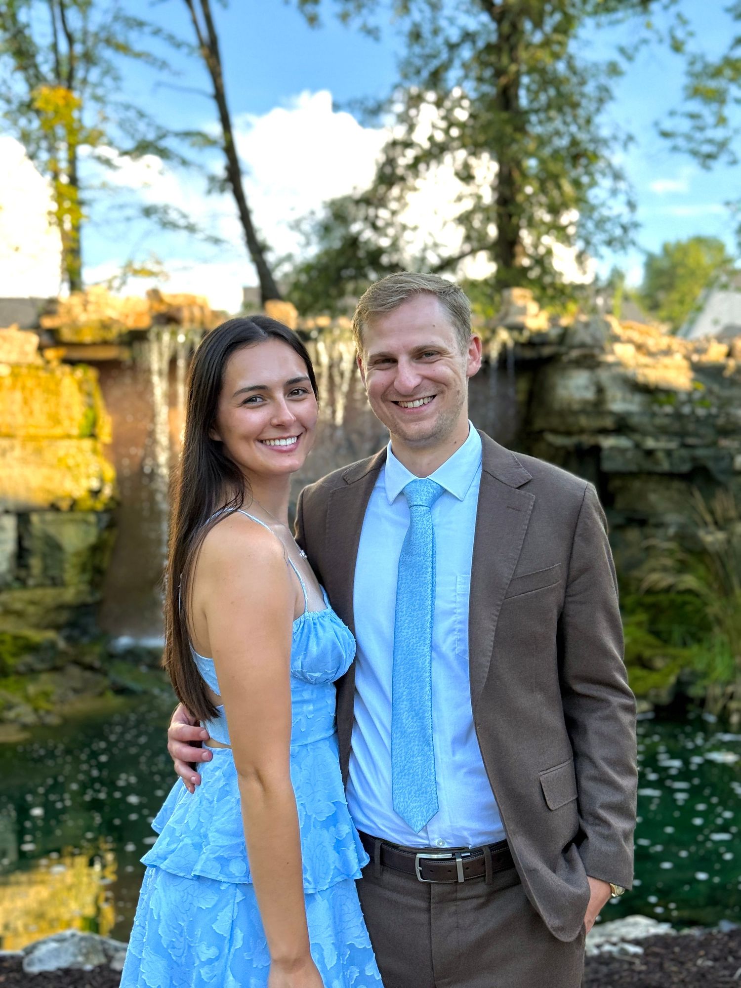 Couple poses together in formal attire by a garden pond with blooming white flowers in the background.