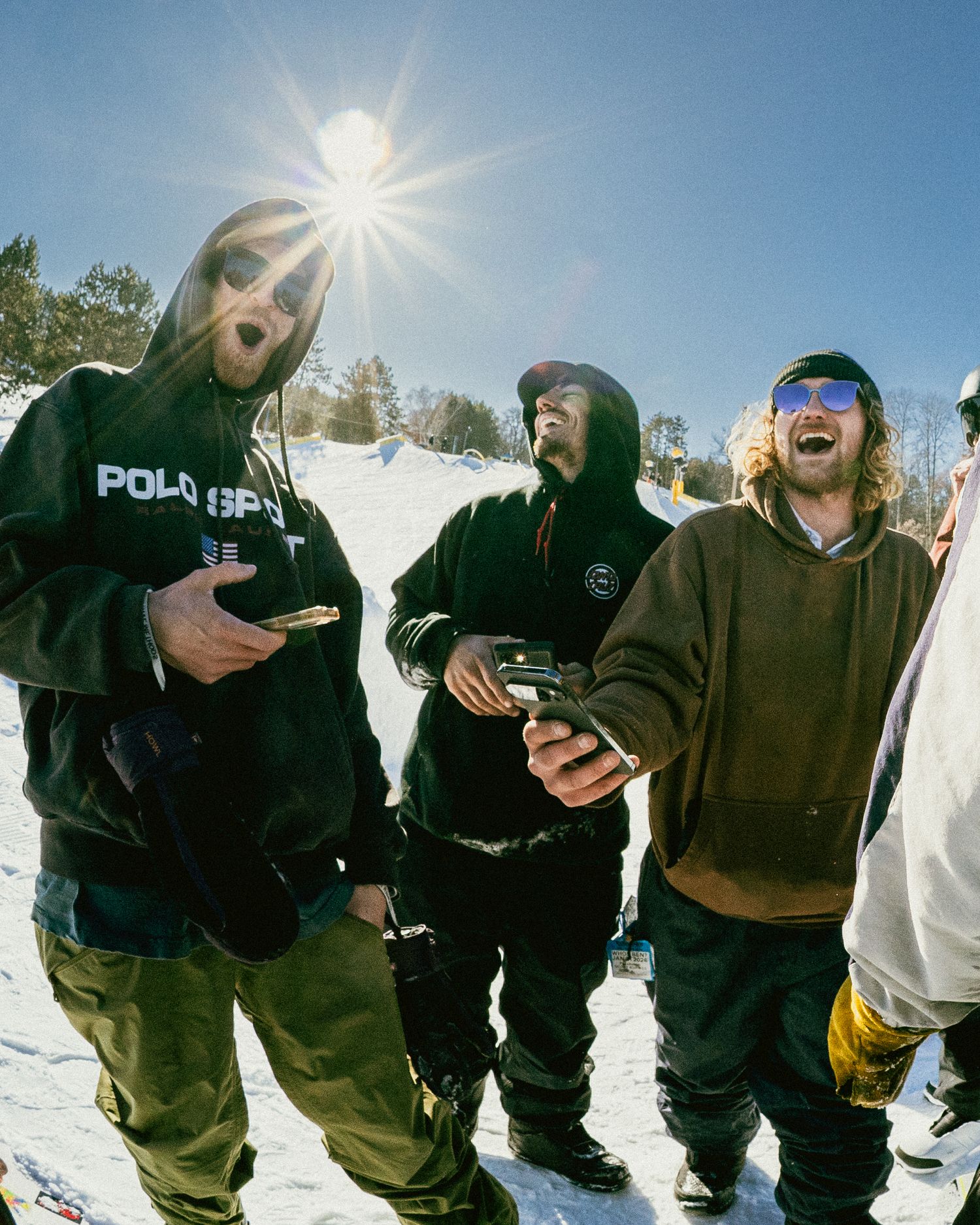 Three friends laughing and holding phones on a sunny snow-covered ski slope wearing winter gear.