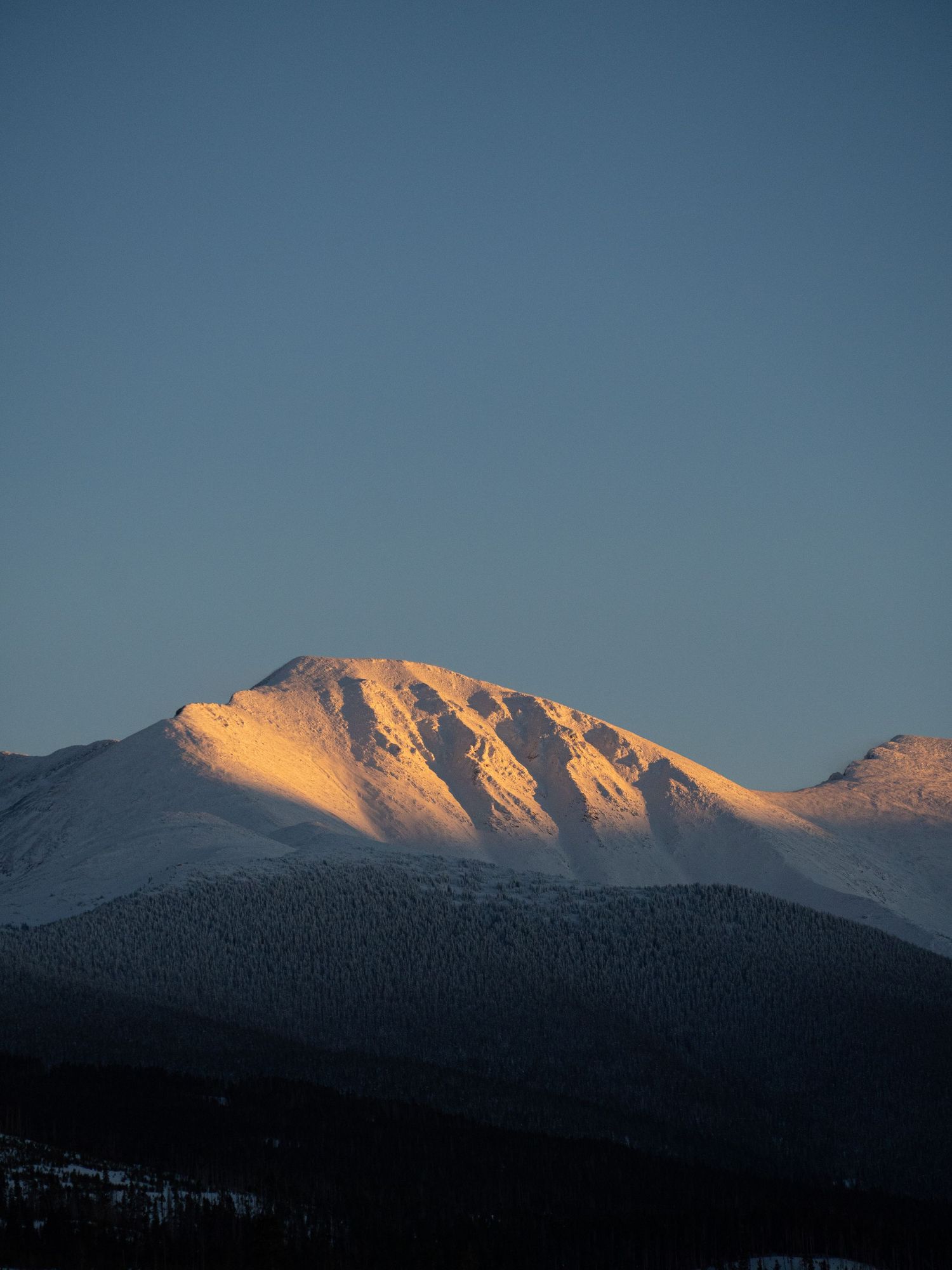 Sunlit snow-capped mountain peak glows golden against deep blue sky at sunset.