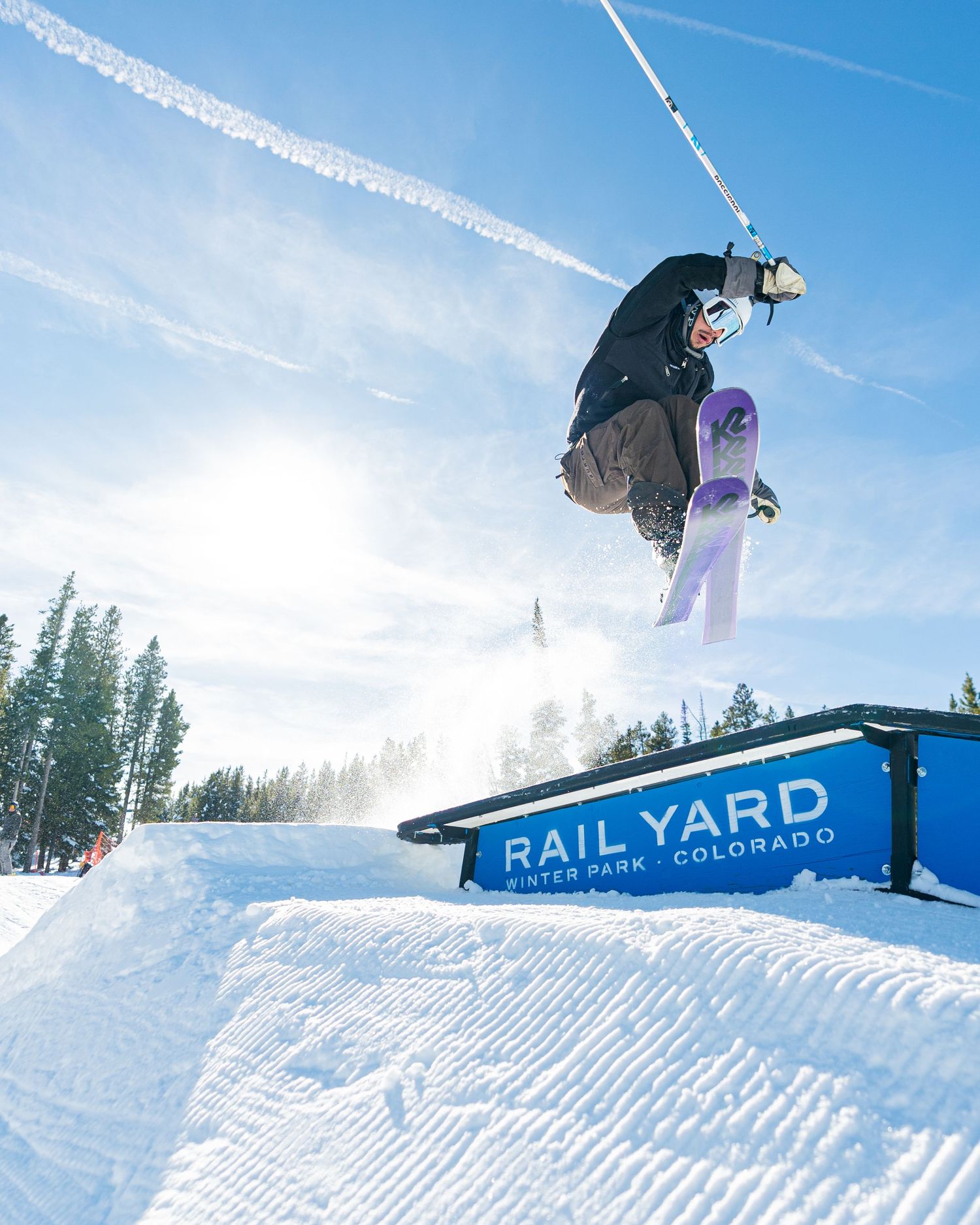 Skier performing aerial trick above snow terrain park rail with blue sky and forest background.