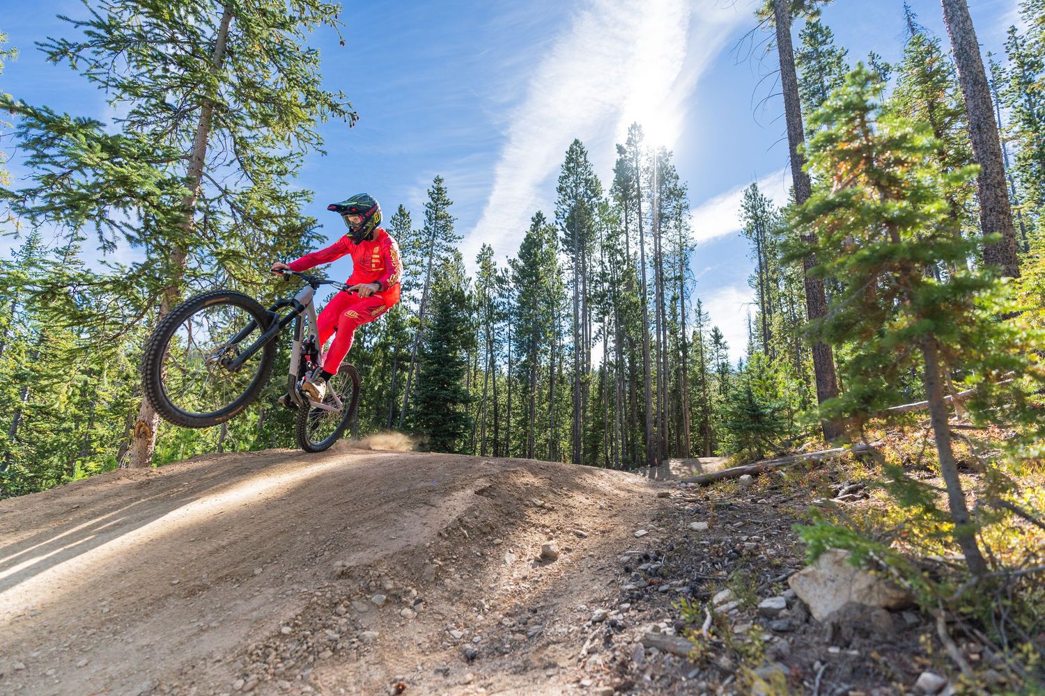 Mountain biker in red gear launches off dirt jump among towering pine trees.