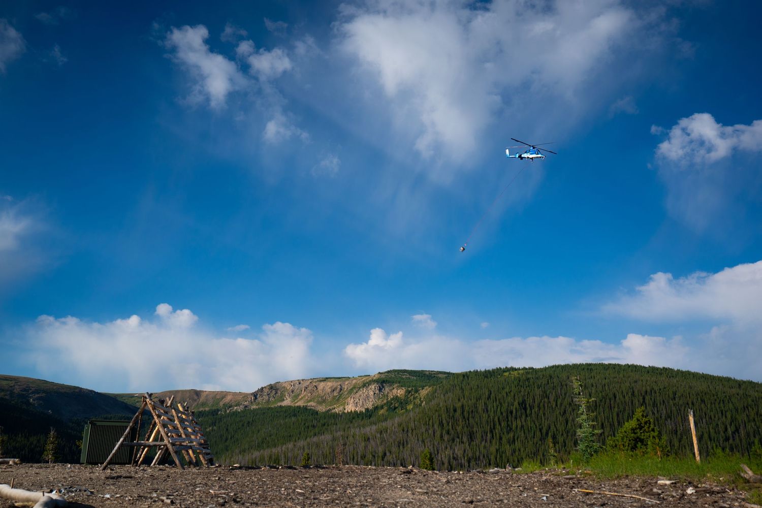 Helicopter flies over mountainous terrain with evergreen forest and wooden structure visible below.