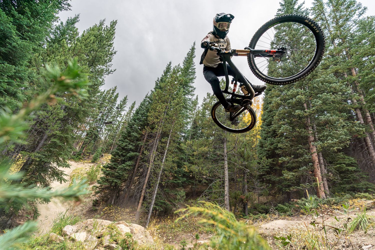 Mountain biker performs aerial trick jump with bike against evergreen forest backdrop.