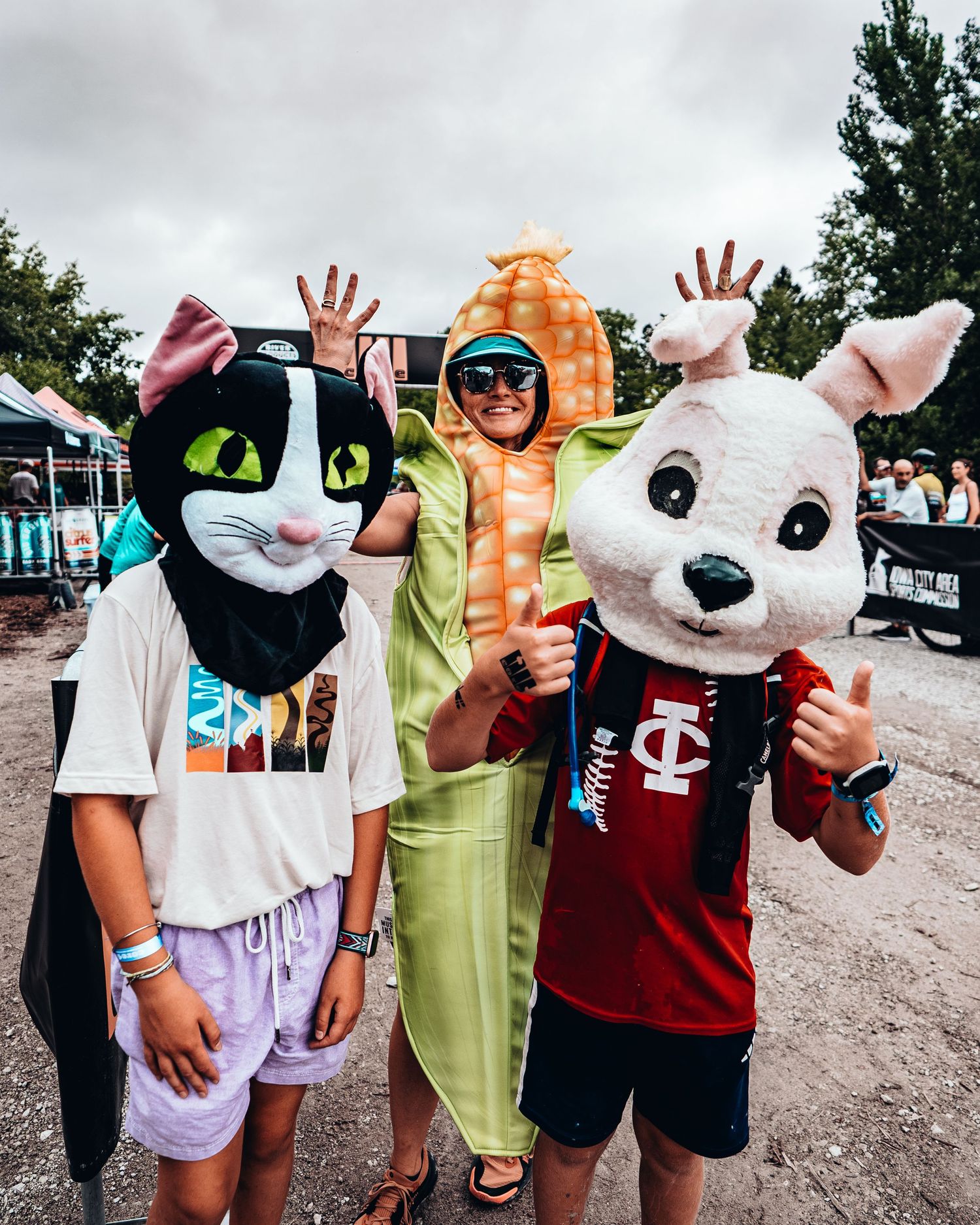 Three people in fun costumes including a cat mask, vegetable outfit, and bunny head at an outdoor festival.