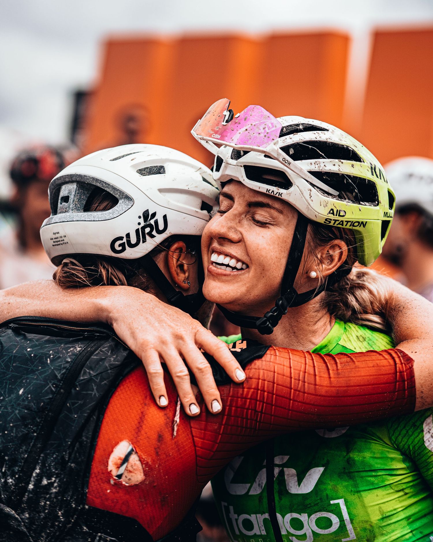Two female cyclists in helmets share a warm embrace after a muddy race, wearing Guru and Liv team gear.