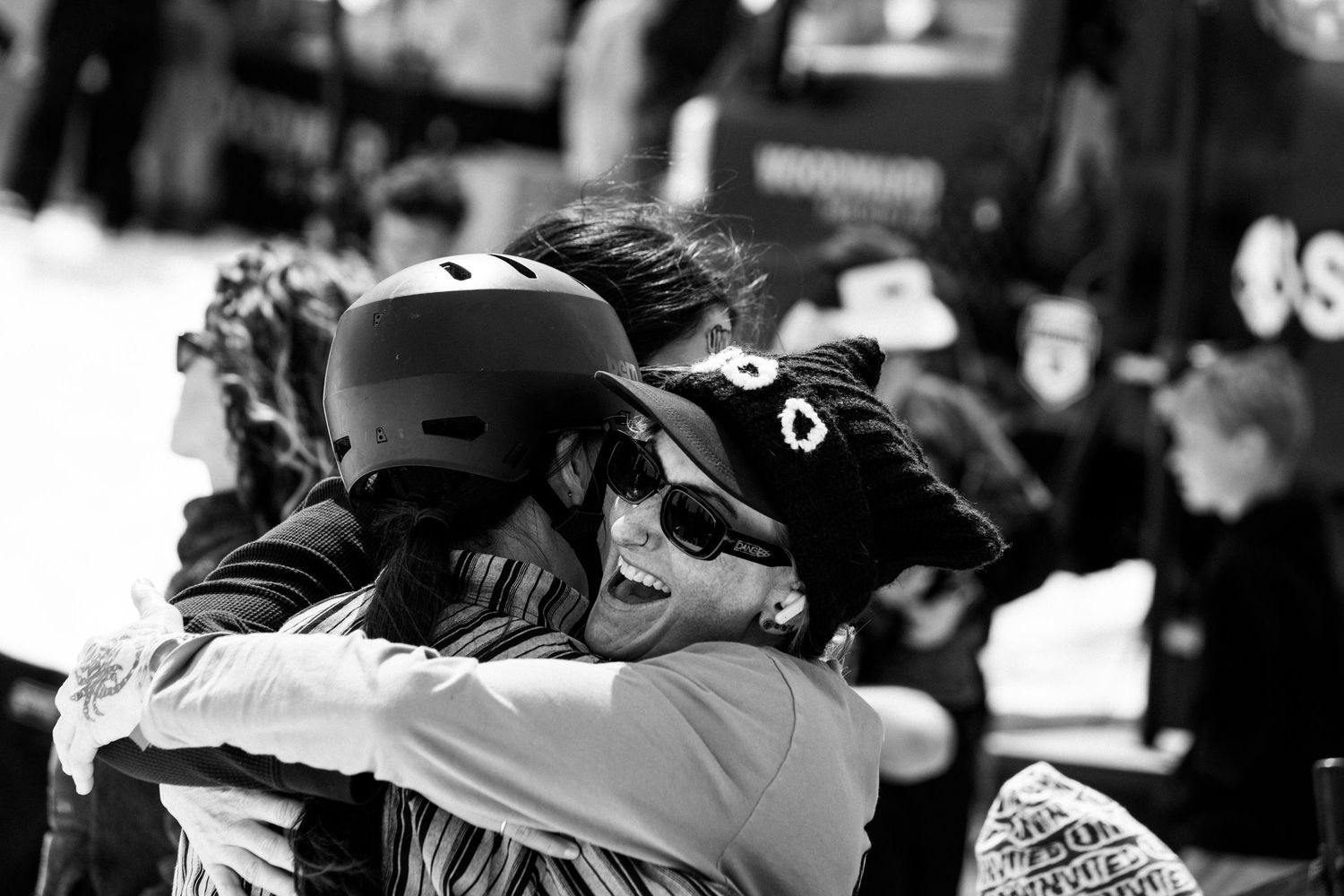 Two skiers in helmets share a warm embrace at a snowy winter sports event in black and white.
