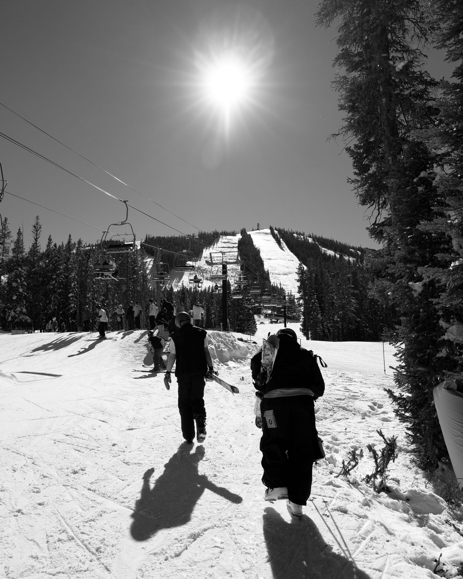 Black and white photo of skiers walking under bright sun with ski lifts and slopes in background.