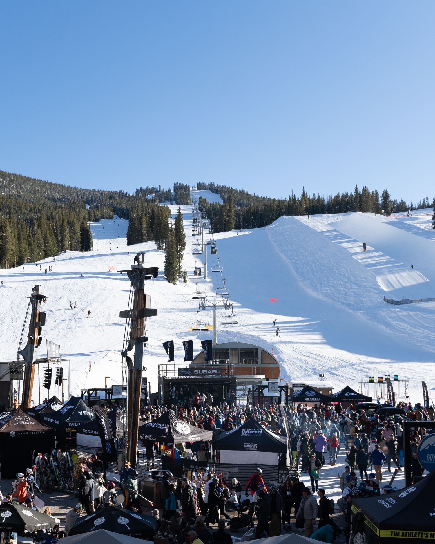 Crowded ski resort base area with chairlifts and snow-covered slopes under blue sky.