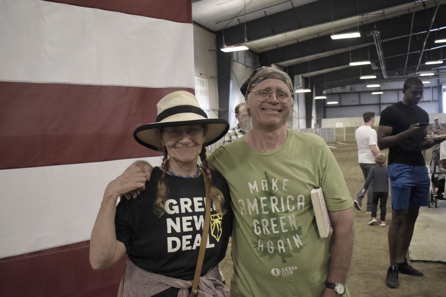 Two supporters wearing campaign-style t-shirts smile together in front of an American flag backdrop at political event.