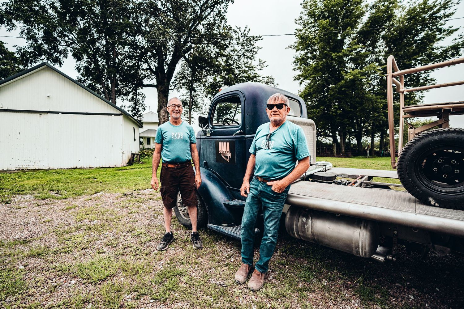 Two men in matching teal shirts stand beside a vintage flatbed truck on a rural farm property.