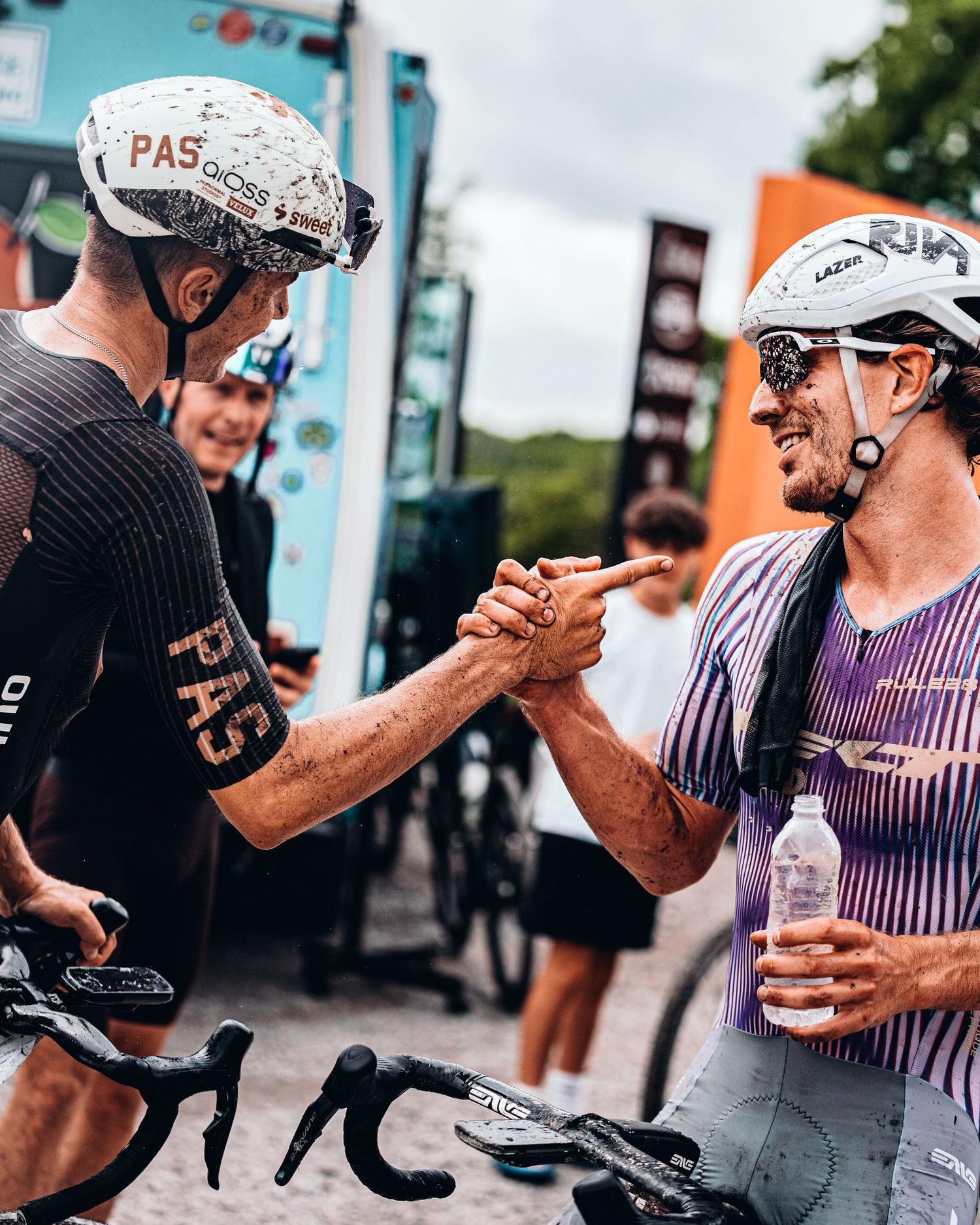 Two cyclists in helmets and jerseys fist bump after a race, one holding a water bottle outdoors.