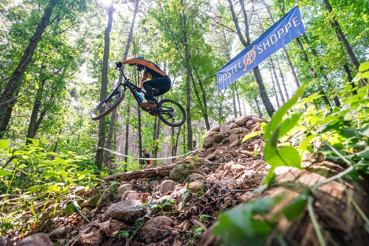 Mountain biker performs an aerial jump on a dirt trail through lush green forest terrain.