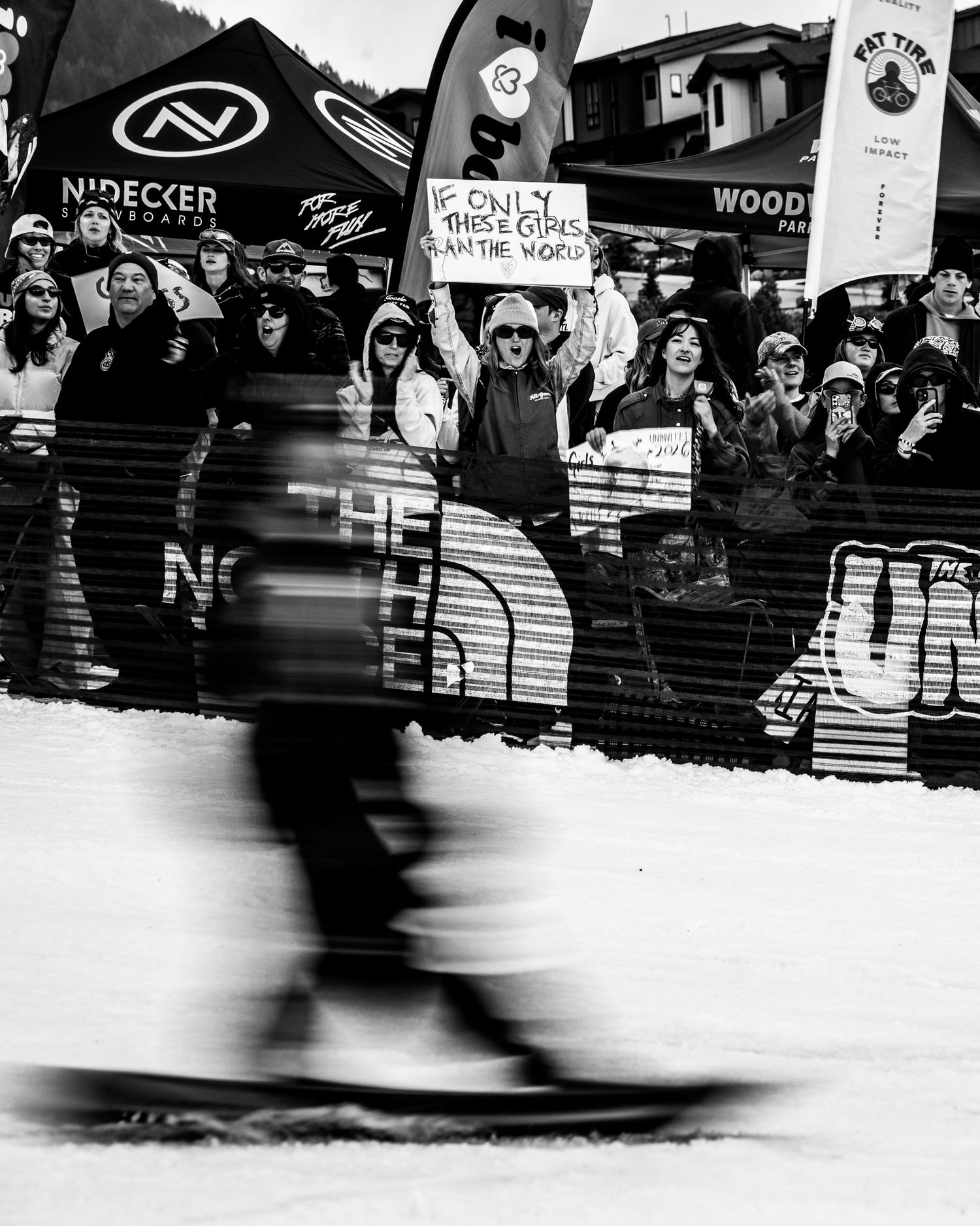 Blurred snowboarder races past cheering crowd holding signs at The Uninvited Invitational competition.