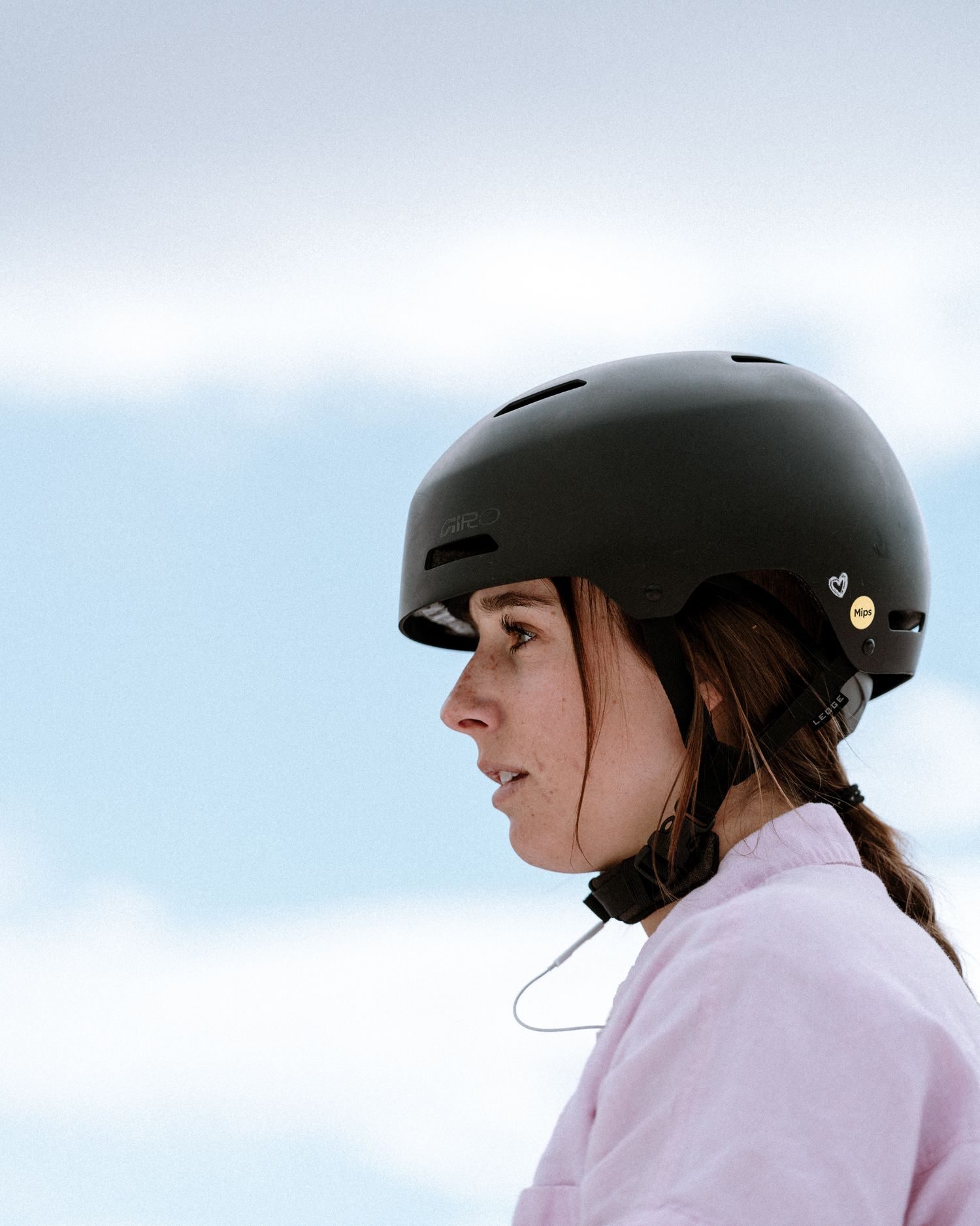 A young woman wearing a black helmet and pink jacket poses against a winter sky backdrop.