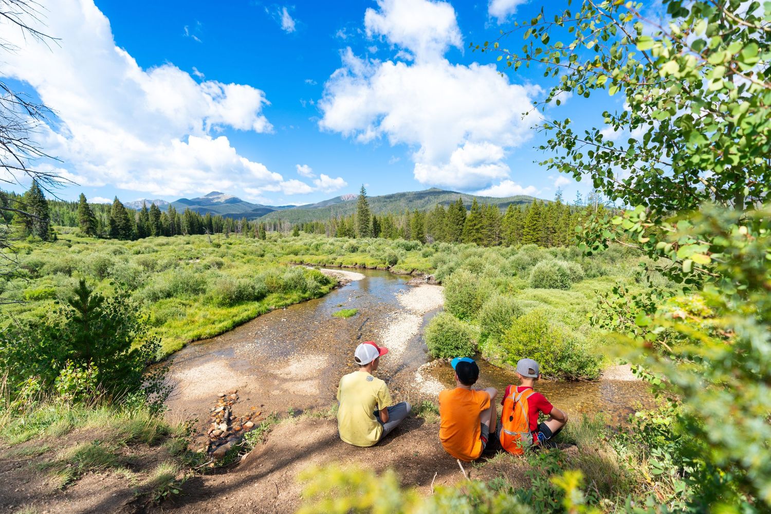 Friends sitting by mountain stream enjoying scenic mountain views on sunny day.