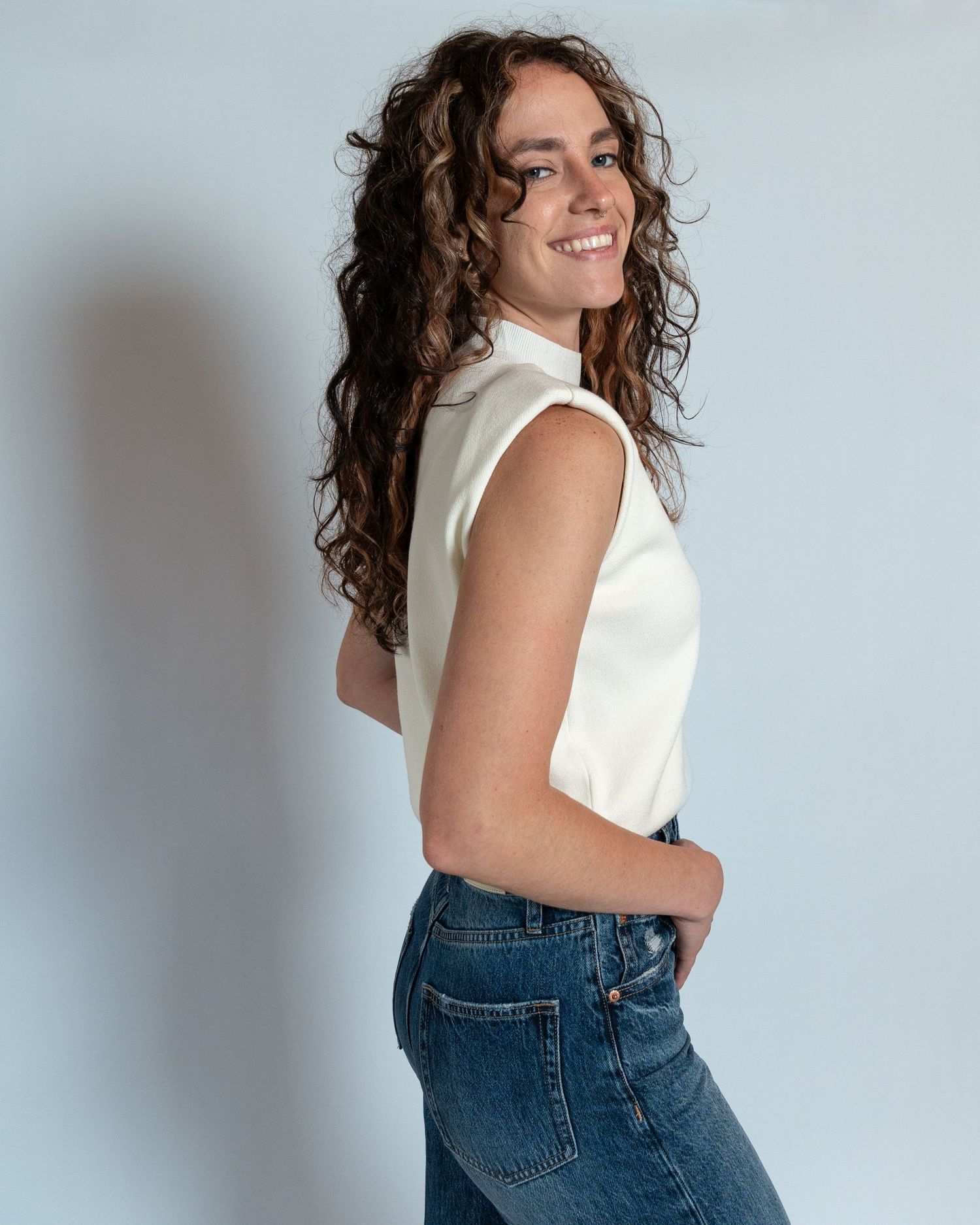 A person in a white sleeveless top and blue jeans poses against a light background while smiling.