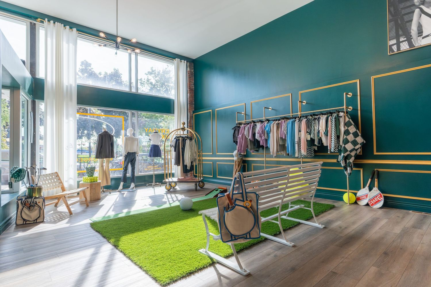 Stylish boutique clothing store with teal walls, gold accents, hanging racks, and green turf rug near large windows.