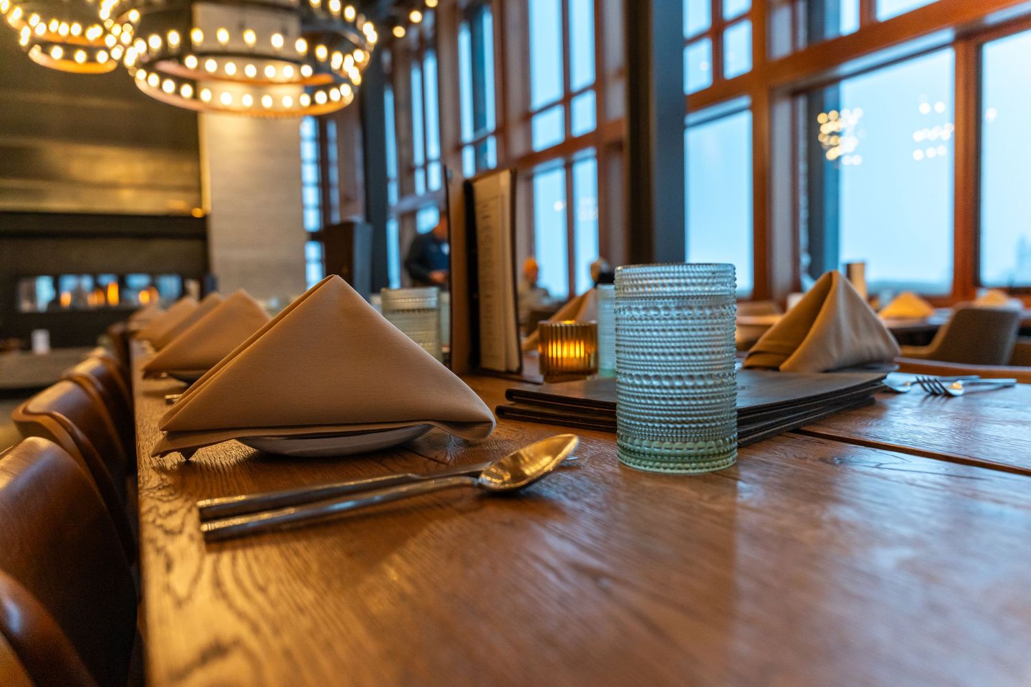 Elegant table setting in restaurant with folded napkins, glassware and warm lighting from circular chandelier above.