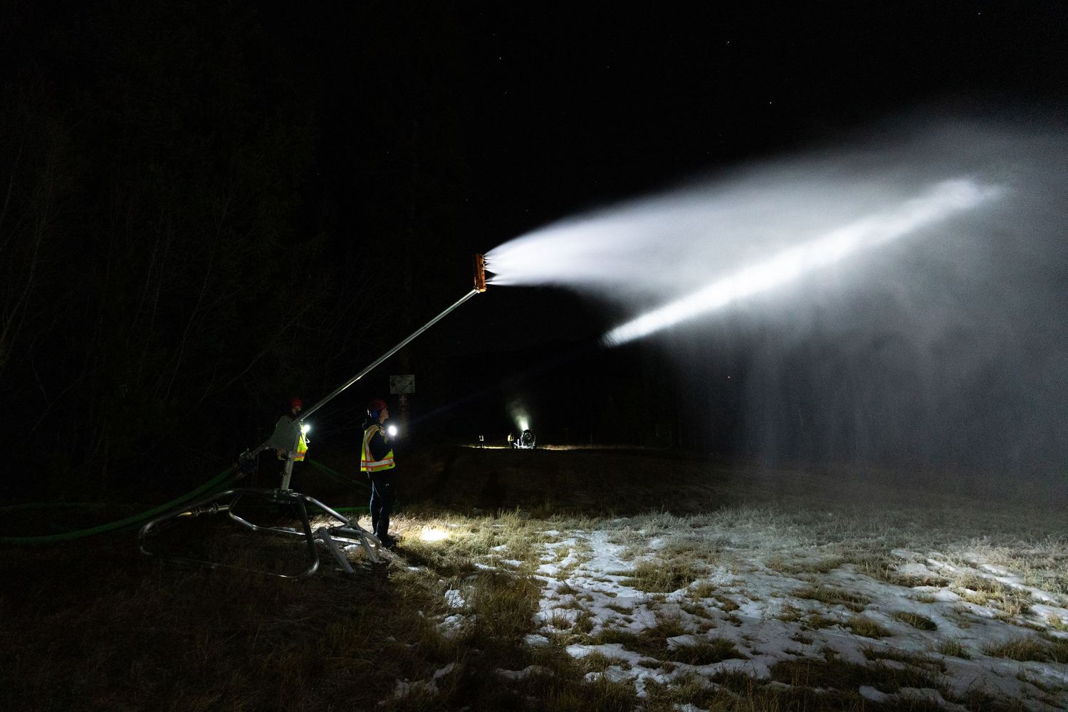 Snow-making equipment sprays artificial snow at night on a ski slope with bright spotlights illuminating the scene.