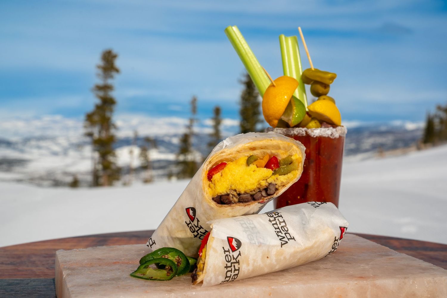 Food and drinks arranged on a wooden table with a snowy mountain backdrop.