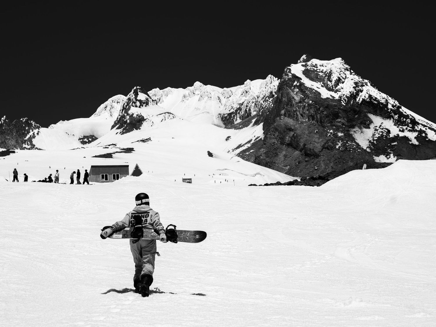 Black and white landscape of snow-covered mountain peak with tiny skiers visible at base.